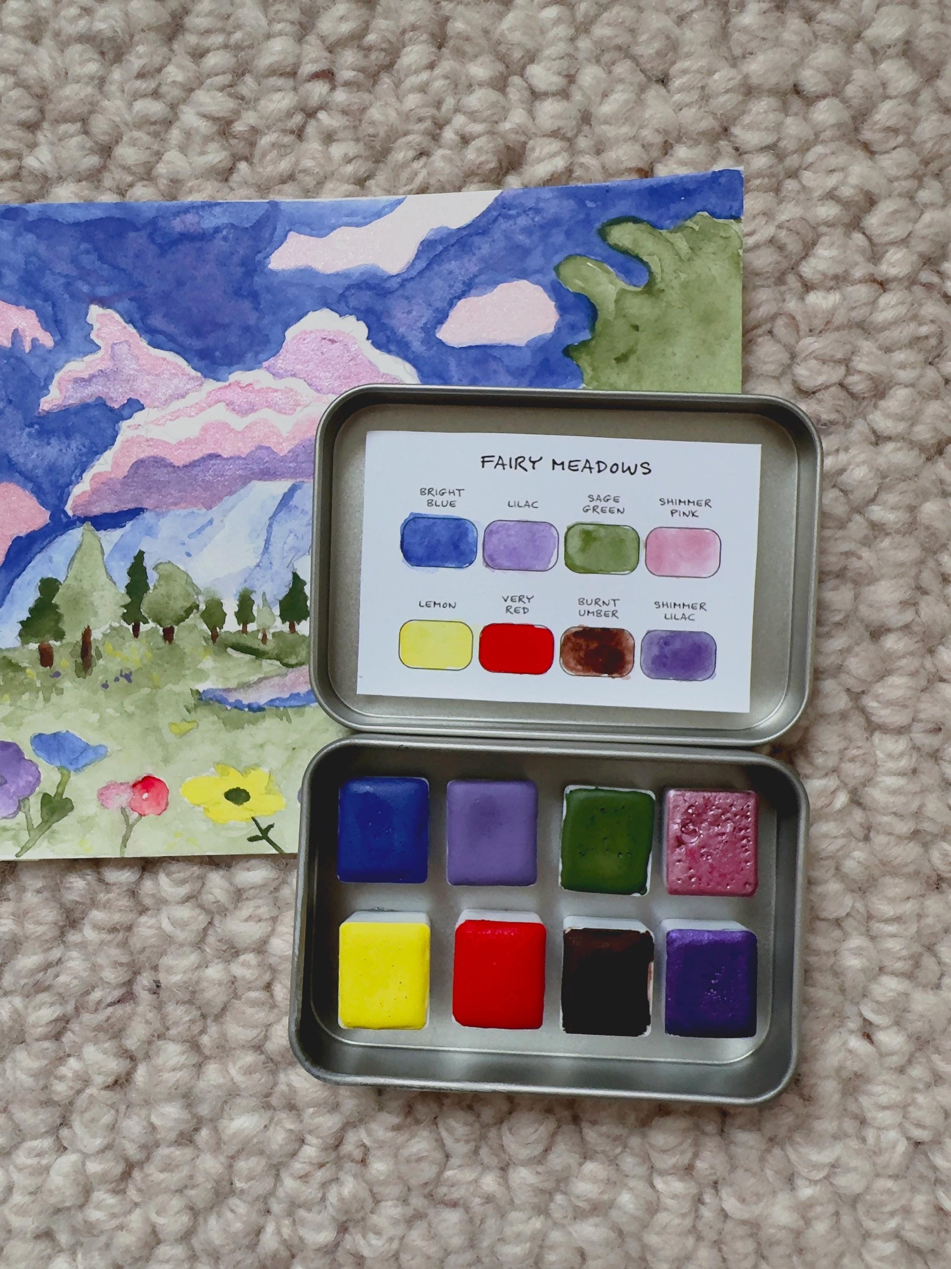 Metallic watercolor paint set with color swatches next to a painted landscape on a carpeted floor.