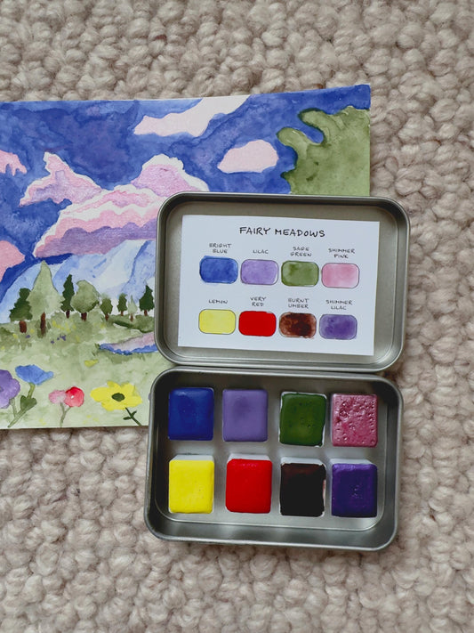 Metallic watercolor paint set with color swatches next to a painted landscape on a carpeted floor.