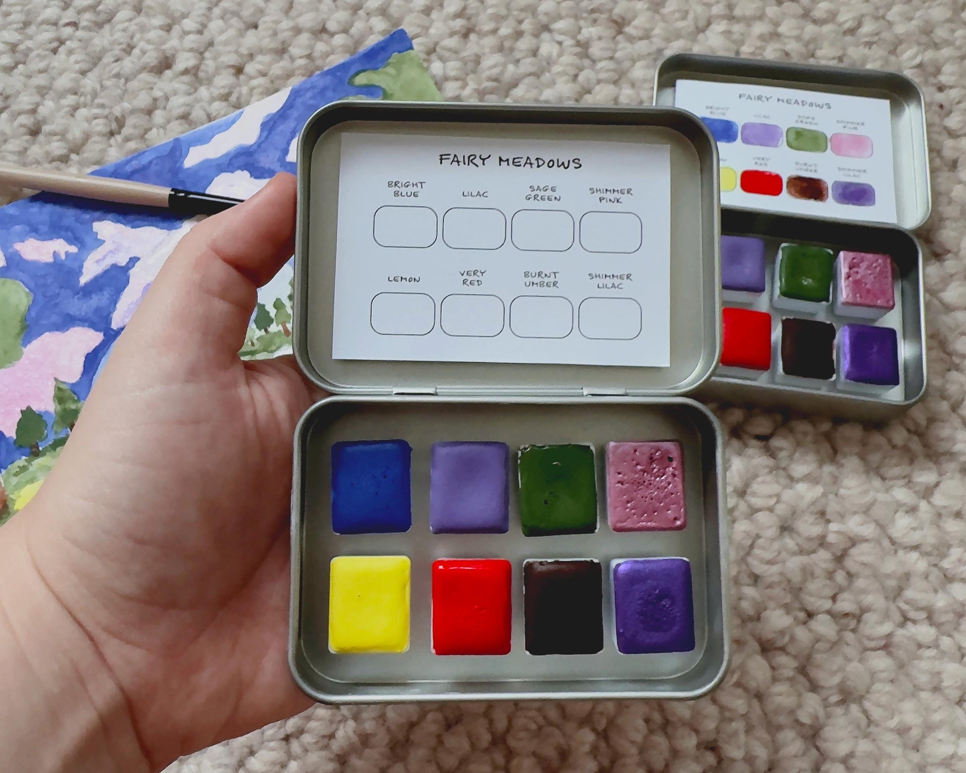 Metal watercolor paint set with colorful swatches held by a hand on a textured surface.